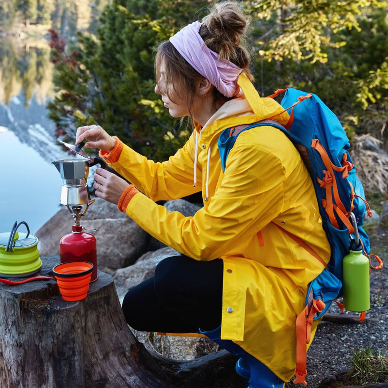 Person in yellow jacket and blue backpack preparing coffee outdoors by a lake